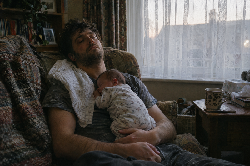 A new dad asleep upright on the sofa at dawn with his newborn asleep on his chest